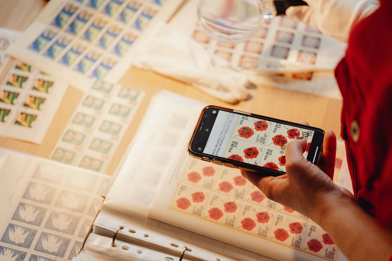 A person uses a smartphone to photograph a collection of colorful postage stamps on a wooden surface.