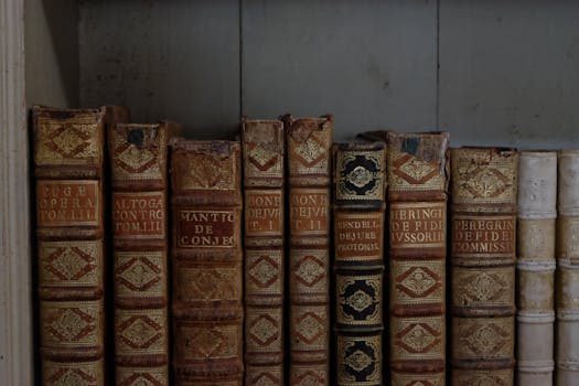 A collection of rare first edition books arranged on a wooden bookshelf, some with dust jackets in protective covers.